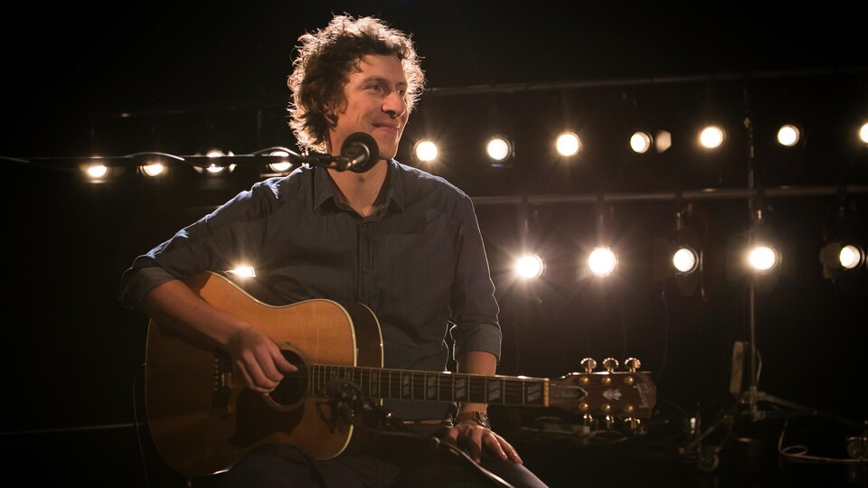 Patrice Michaud assis sur un siège sur scène, avec sa guitare.
