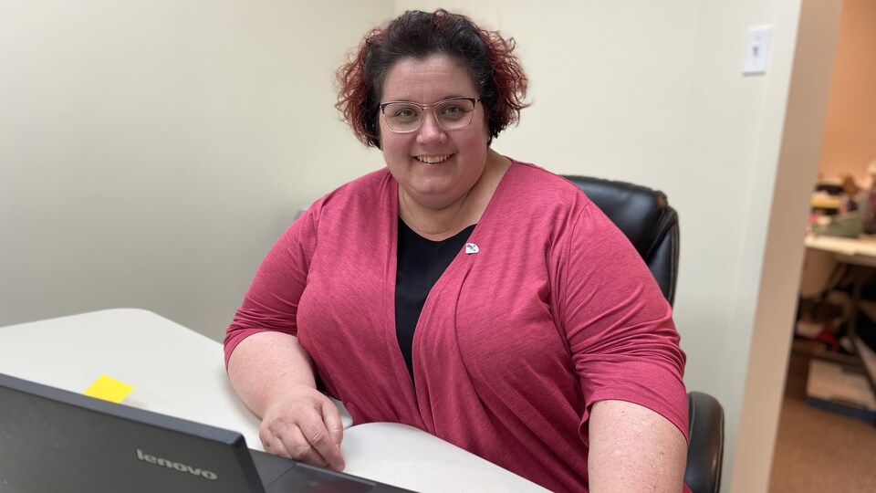 Une femme travaillant installée derrière un bureau sur lequel est posé un ordinateur portable et un téléphone cellulaire sourit à la caméra.