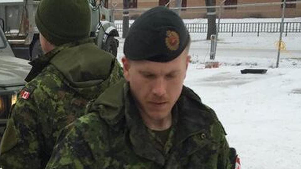 Un homme vu en gros plan. Il est revêtu de l'uniforme de l'armée. La photo a été prise à l'extérieur, en hiver. L'arrière-plan montre d'autres soldats et des véhicules de l'armée. 