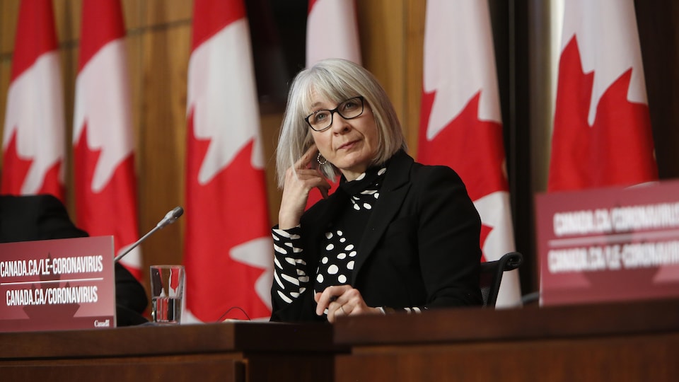 La ministre écoute une question lors d'une conférence de presse, assise devant des drapeaux canadiens. 