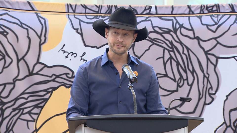 Le chanteur Paul Brandt fait une allocution devant une estrade avec micro. Derrière lui, une grande toile avec le logo de sa campagne de sensibilisation (des roses) est affichée.