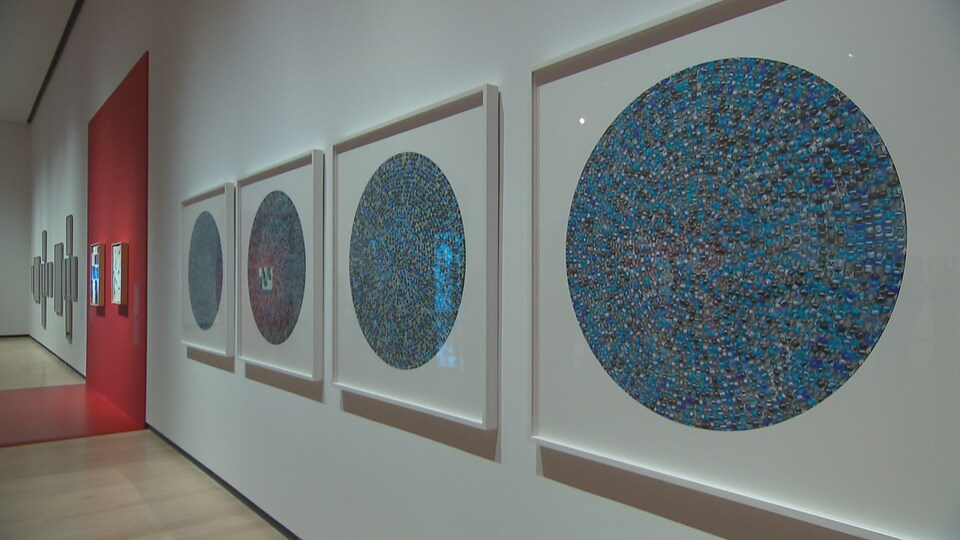 Quatre tableaux sont accrochés au mur. Il y a un cercle de petites billes au centre de chaque carré. D'autres œuvres sont placées plus loin sur le mur. 