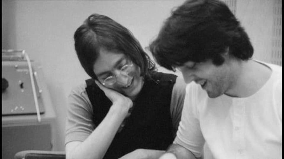 John Lennon est assis à côté de Paul McCartney, qui écrit.