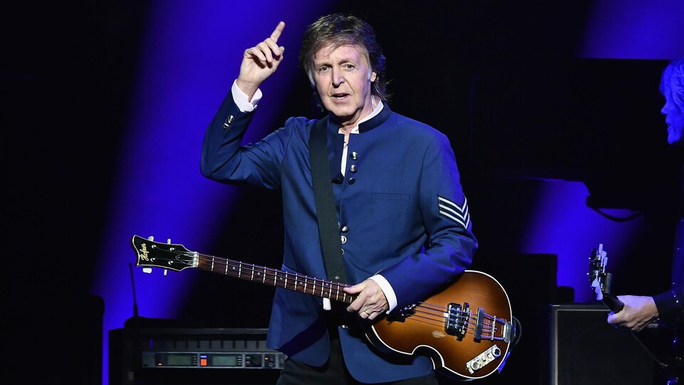 Paul McCartney sur scène, le bras droit levé, avec une guitare basse.
