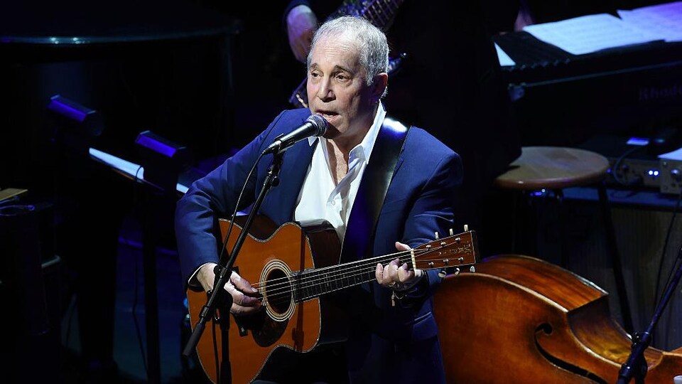 Paul Simon porte un costume bleu et tient une guitare sur scène. 