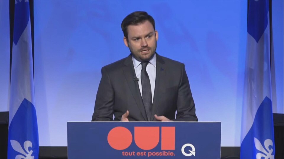Un homme parle devant un lutrin avec le logo du PQ où il est écrit : « Oui, tout est possible ».