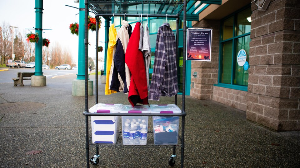 Une station de provisions, sur laquelle sont accrochés des vêtements sur des cintres et, près du sol, des boîtes de rangement contenant des bouteilles d'eau et des produits d'hygiène.
