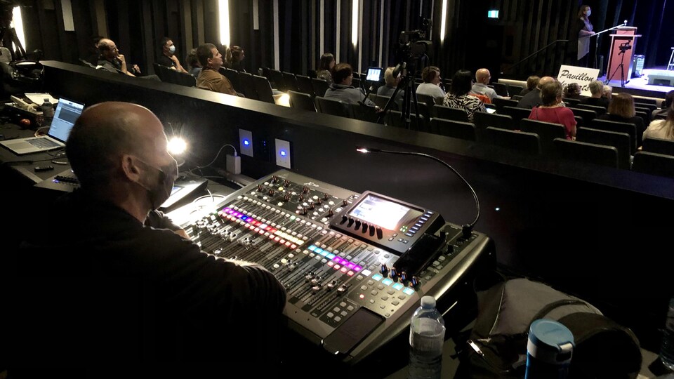 Dans la salle de spectacle. Un homme est assis devant une console de son. En arrière-plan on aperçoit des spectateurs assis et une personne sur la scène.