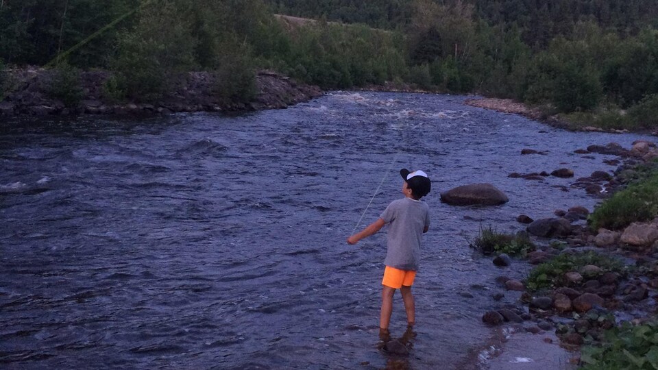Un jeune garçon pêche à la mouche dans une rivière calme entourée de forêt.