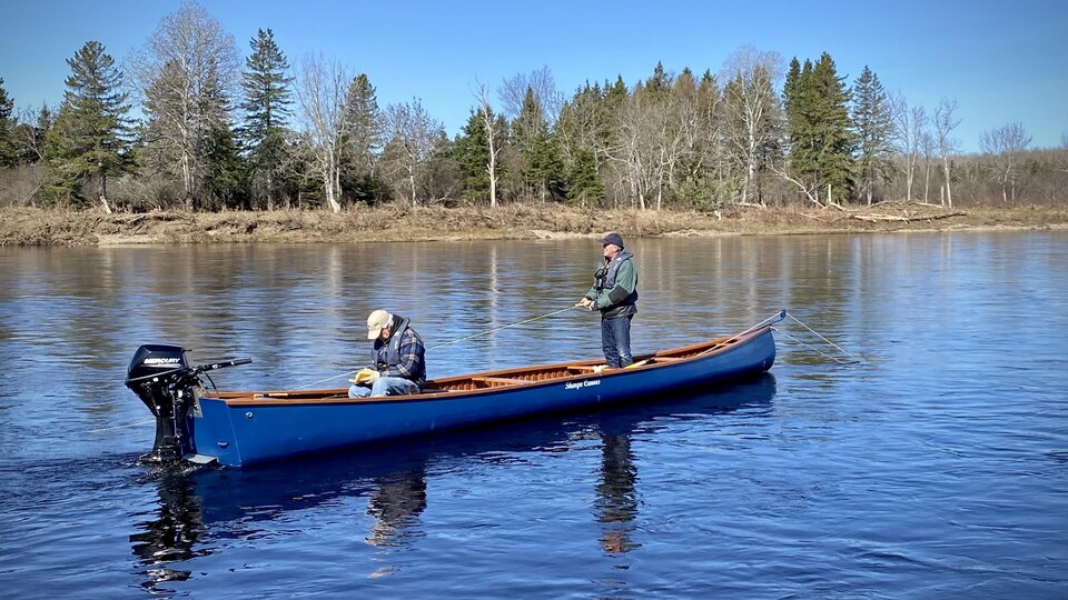 Deux pêcheurs amateurs sont à bord d'un canot à moteur sur la rivière Miramichi. Pendant qu'un, debout, surveille sa ligne à l'eau. L'autre, assis, prépare son équipement.