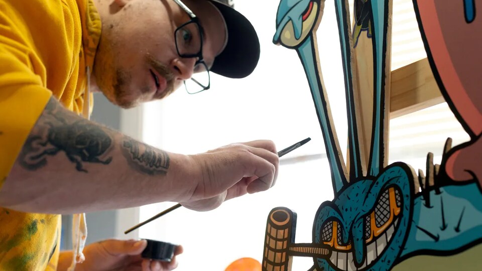 L'artiste Daarian Woroniak tient un pinceau dans sa main droite et s'apprête à colorer son oeuvre sur bois.
