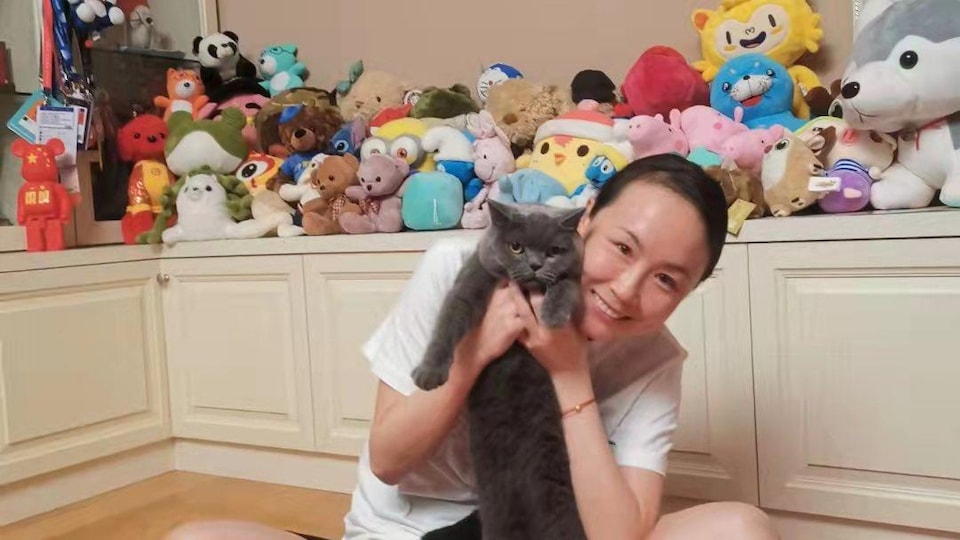 Une femme tient un chat en souriant à la caméra dans une pièce remplie de peluches. 