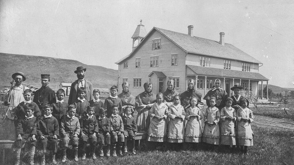 Photo d'archives où l'on voit des élèves et leurs superviseurs regroupés sur le terrain devant l'édifice. Les garçons sont à gauche, les filles à droite.