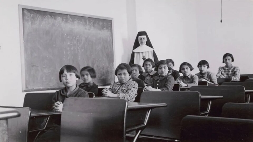 Une photo noire et blanc d'un groupe d'élèves et une religieuse dans une salle de classe du pensionnat de Pimicikamak en 1940.