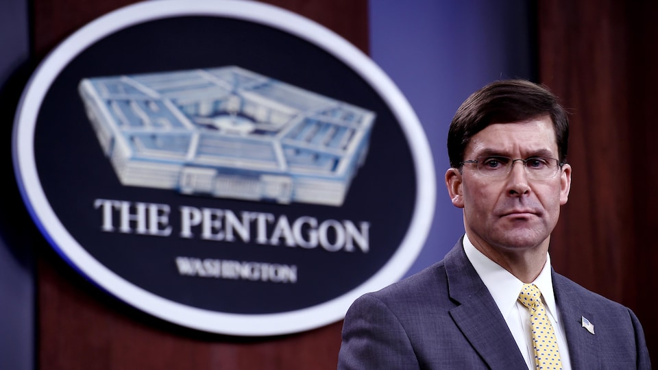 Un homme en complet affiche un air inquiet. Derrière lui se trouve un logo du Pentagone.