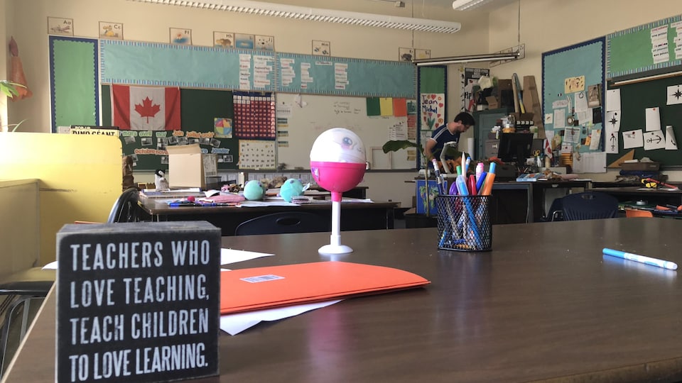 Une table dans une classe avec des objets dessus et un enseignant à son bureau.