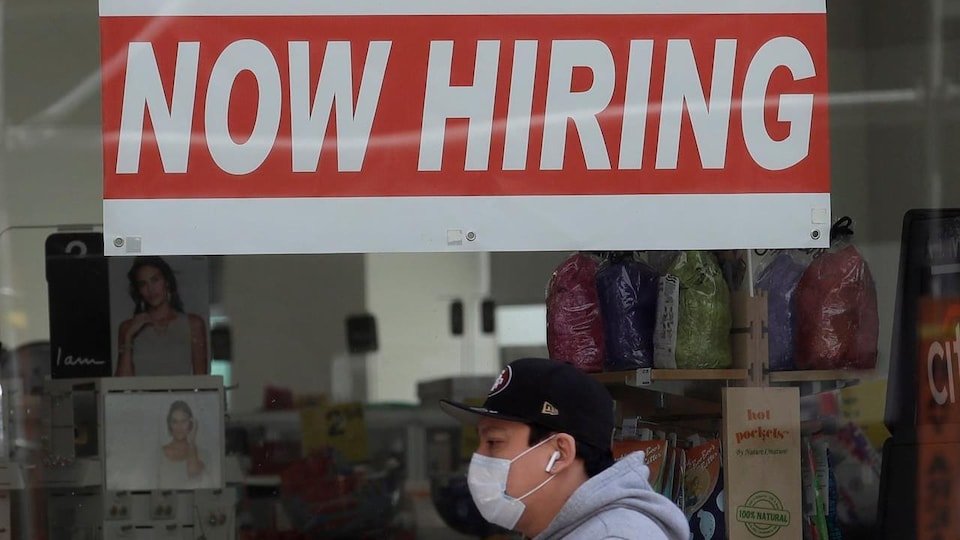 Un homme passe devant un magasin. Il porte un masque et des écouteurs sans fil. Dans la vitrine, une affiche dit en anglais : Nous embauchons".