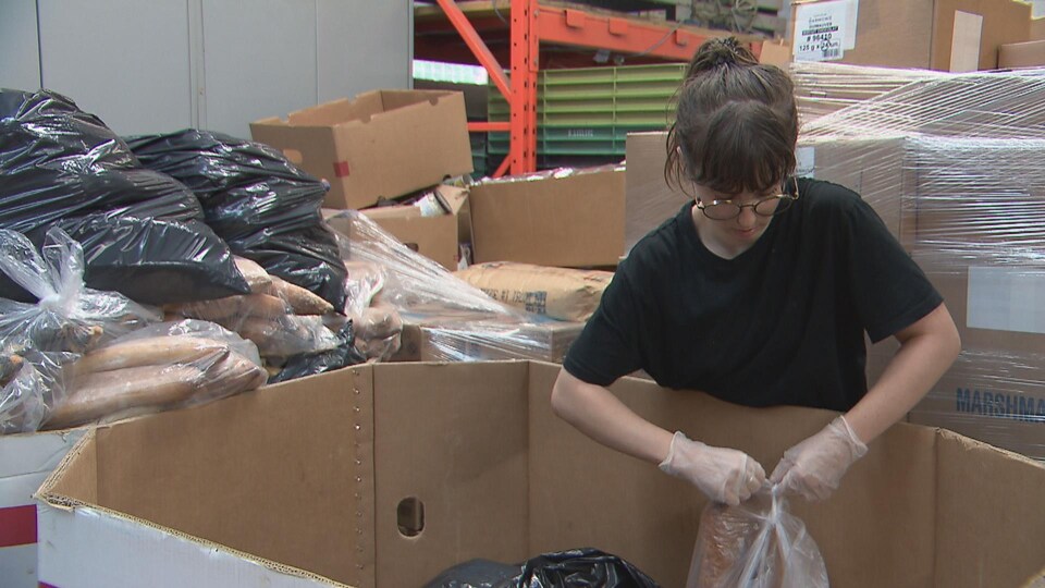 Une femme est en train de fermer un sac de plastique dans un entrepôt. Elle est penchée au-dessus d'un grand carton.