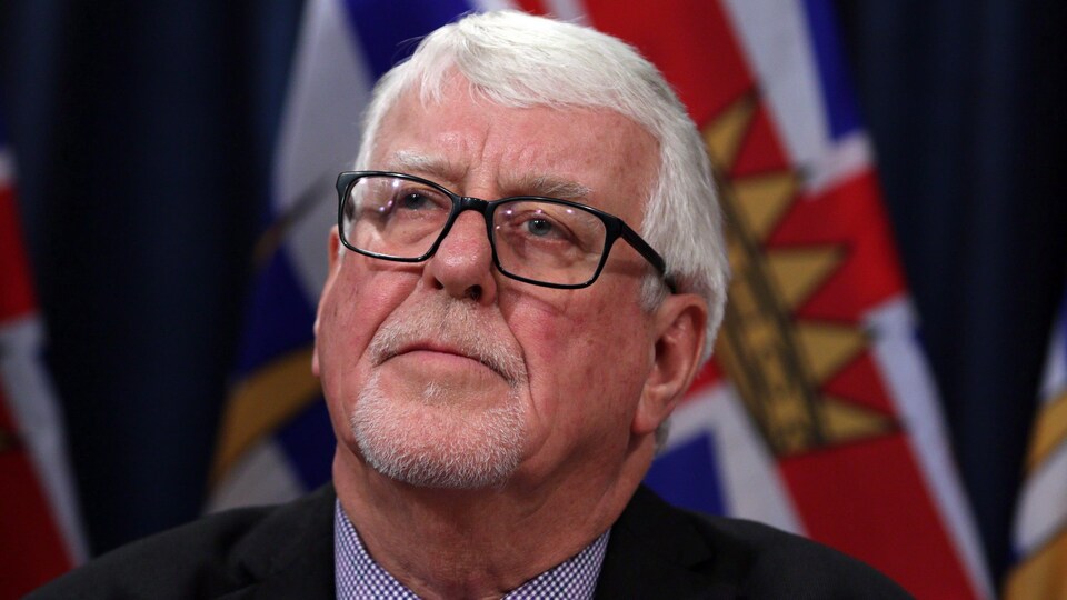 Photo cadrée serrée de la tête et des épaules de Perry Kendall avec en-arrière plan flou le drapeau de la Colombie-Britannique.