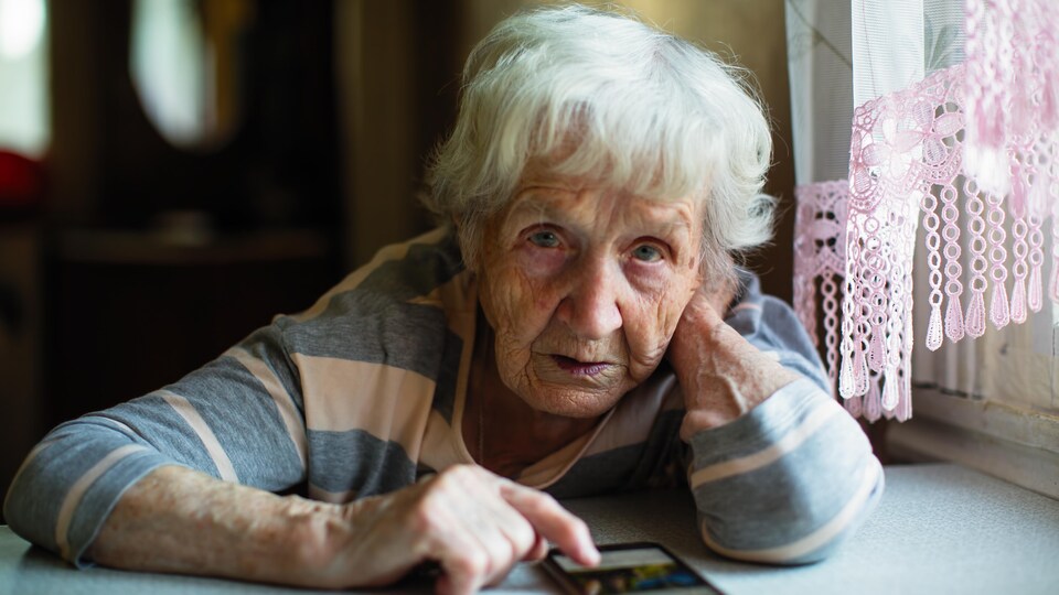 Une grand-mère assise à sa table de cuisine avec un téléphone cellulaire posé devant elle.