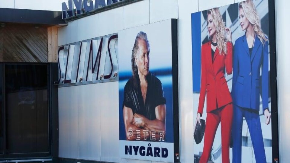 Une grande affiche murale sur laquelle on voit à gauche de l’image un homme et à droite deux dames debout en tailleur.