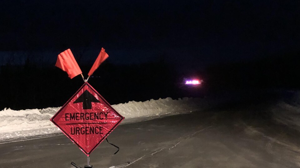 Un panneau sur lequel est écrit URGENCE au milieu d'une autoroute bloqué. Au loin, les gyrophares d'une voiture de police.
