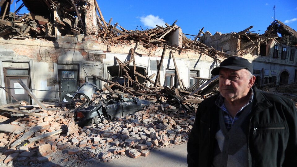 Un homme est posté en face d'un long bâtiment en ruines, devant lequel une voiture est couverte de débris.
