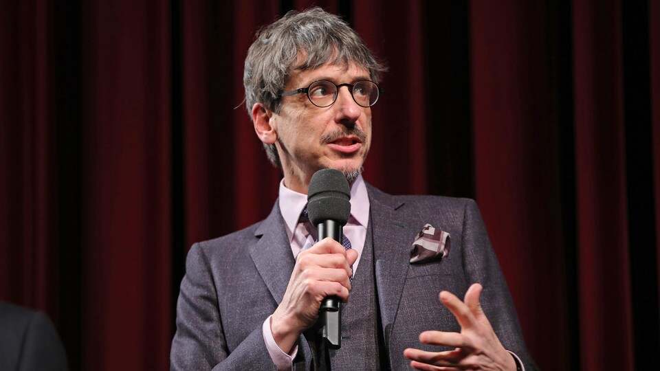 L'homme porte des lunettes et parle dans un micro.