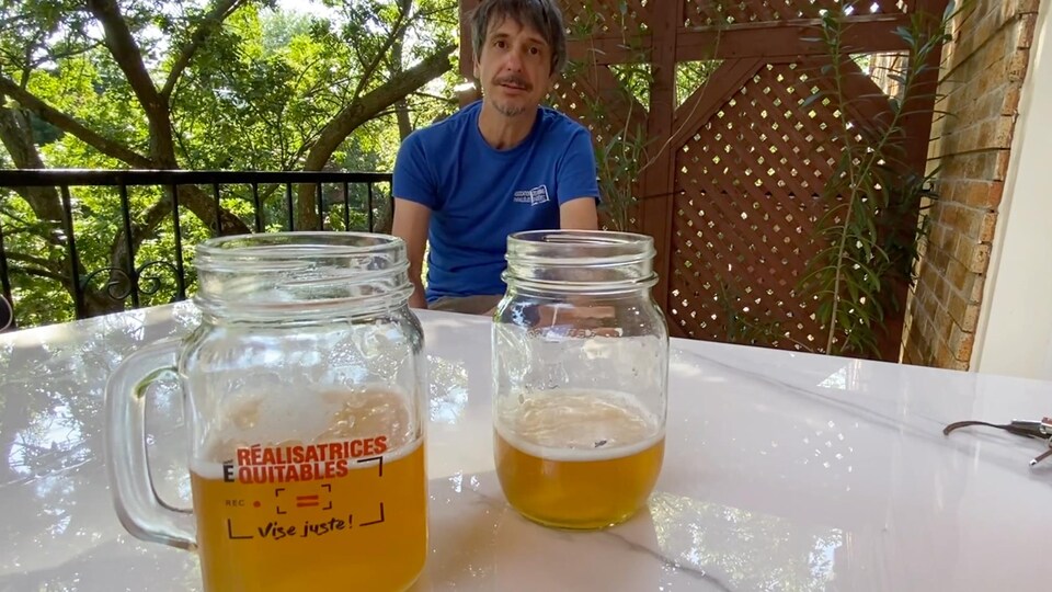 L'homme est assis et deux pots Mason contenant de la bière à moitié sont posés sur une table devant lui. Un des pots porte le logo de la campagne de Réalisatrices équitables.
