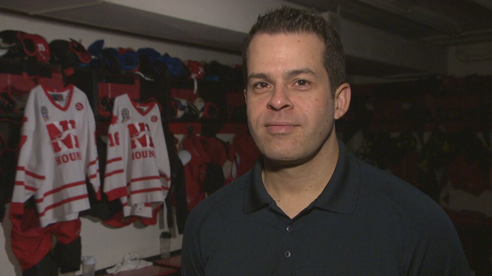 Un homme avec un chandail gris pose devant la caméra, le regard fier. Il est debut dans un vestiaire de hockey et on voit les chandails des Houds de Notre Dame en arrière-plan.
