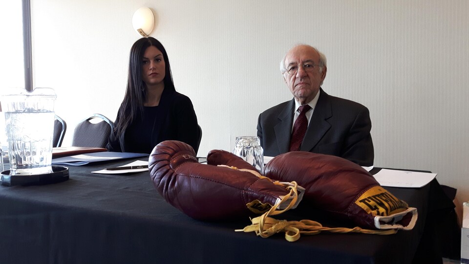 Philippe Sauvageau et son avocate sont assis derrière une table en attendant le début d'une conférence de presse. Une paire de gants de boxe est déposée sur la table.