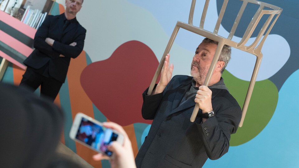 On voit Philippe Starck avec une chaise sur la tête, devant des gens qui le prennent en photo.