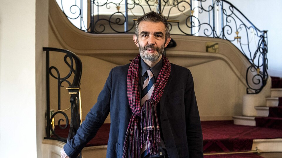 Le journaliste pose devant des escaliers recouverts d'un tapis rouge. Sa barbe poivre et sel cache ses blessures au visage. Il porte un veston bleu et un foulard rouge.