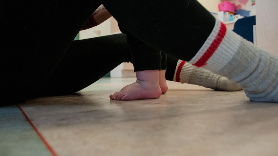 Les petits pieds d'un bébé sont photographiés en gros plan; l'enfant se tient entre les jambes d'un adulte vêtu de pantalons noirs et de chaussettes de laine et qui est assis sur le sol. On ne voit pas les visages.