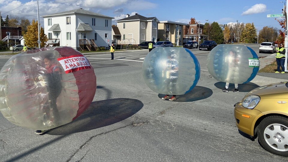 Trois enfants traversent une intersection dans des bulles gonflables, devant une voiture arrêtée.