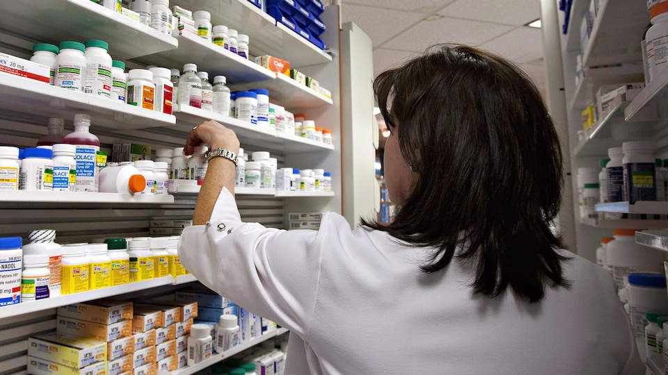 Une technicienne prend une bouteille de pilules sur une des étagères d'une pharmacie.