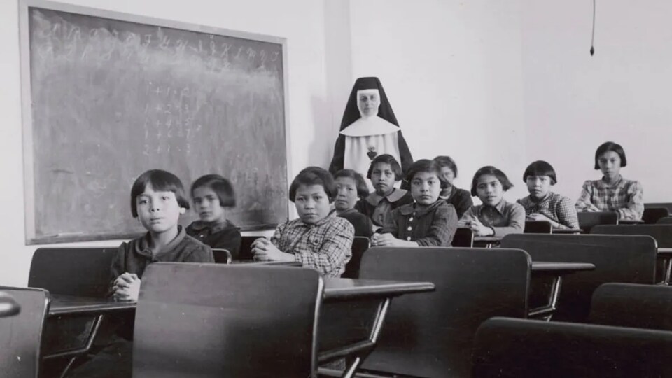 Un groupe d'élèves assis dans une salle de classe du pensionnat de Cross Lake et une enseignante religieuse se tient debout.