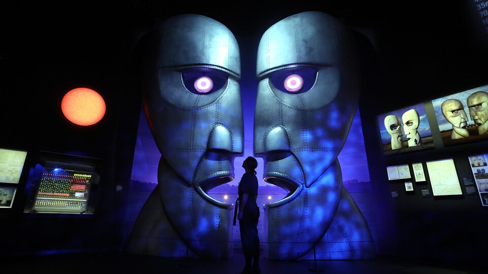 Une femme se tient debout dans une salle d'exposition, devant de grands visages métalliques. 