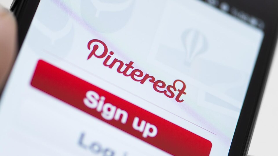 Une photo montrant une main qui tient un téléphone intelligent sur lequel est affiché l'écran d'accueil de l'application Pinterest, avec le logo de l'entreprise et des boutons servant à se connecter.