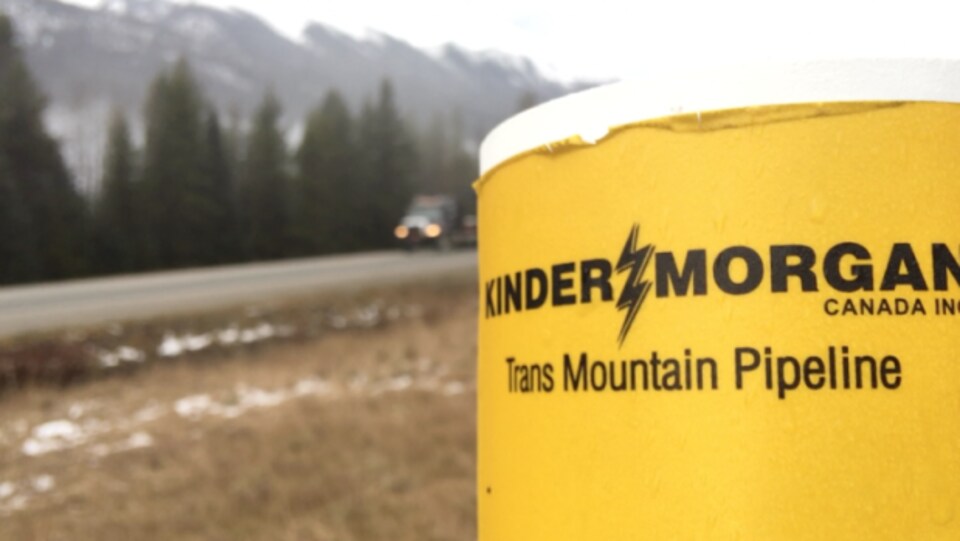 Logo de l'entreprise Kinder Morgan en premier plan, en bordure d'une route. L'entreprise Kinder Morgan affirme que l'attente des permis d'expansion du pipeline TransMountain, en Colombie-Britannique, a des répercussions coûteuses pour le projet.