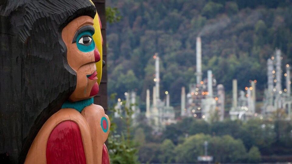 Un totem de profil avec en arrière-plan de la forêt et une usine de pétrole. 