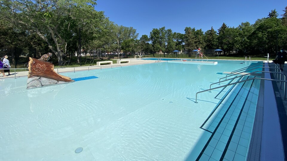 Une piscine extérieure en forme de L avec ses rampes et ses marches. Un sauveteur est assis sur sa chaise et un module de jeu aquatique illustrant un castor sur un tronc d'arbre figure au beau milieu de la piscine.