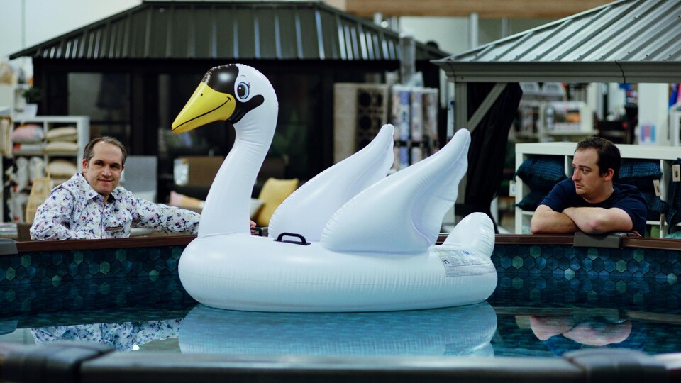 Deux hommes sont accoudés à une piscine hors terre dans laquelle flotte un cygne en plastique.