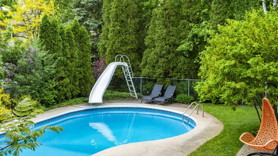 Une piscine avec une glissoire dans un jardin entourée d'arbres matures.