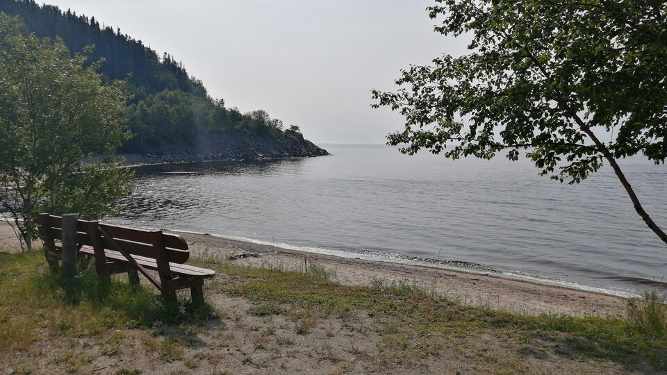Un banc de parc en bois est installé face au Saint-Laurent, en bordure d'une plage de sable.