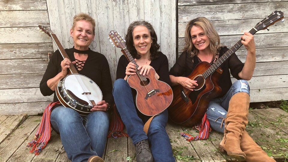 Trois femmes assises devant une grange avec leur instrument de musique à la main.