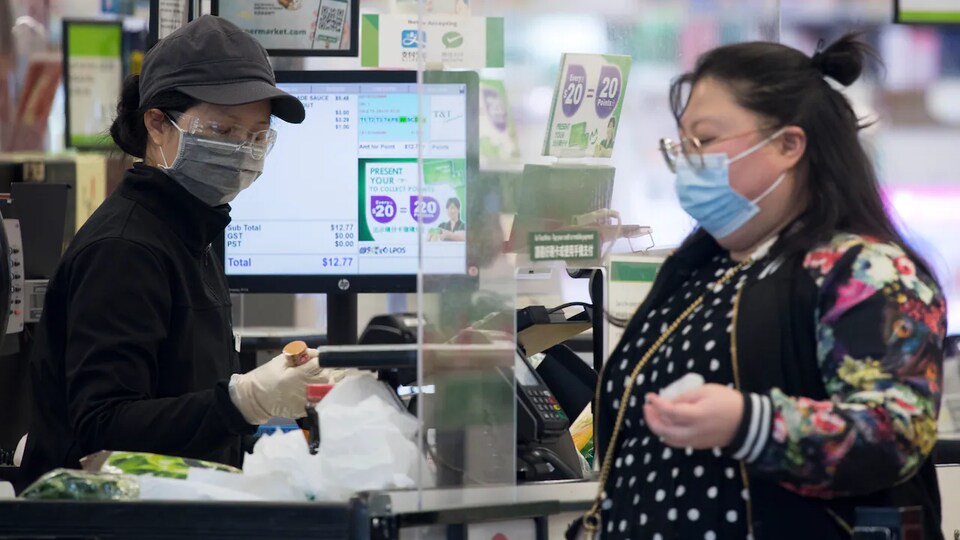 La caissière d'une épicerie et une cliente séparées par une paroi de plexiglas à la caisse.
