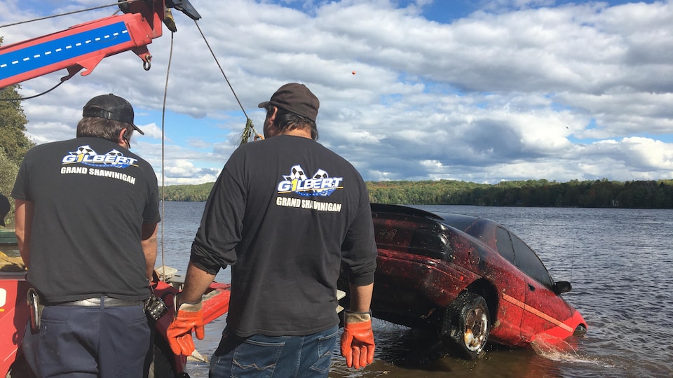 Deux opérateurs de dépanneuse remorquent un véhicule. L'automobile est tirée par le crochet de la dépanneuse et sort de la rivière.