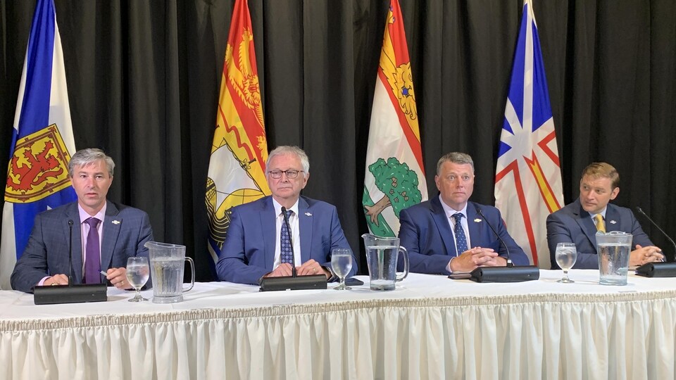 Les quatre messieurs en veston bleus, portant des cravates, sont assis à une même table couverte d'une nappe blanche. Les drapeaux des quatre provinces de l'Atlantique sont plantés derrière eux.