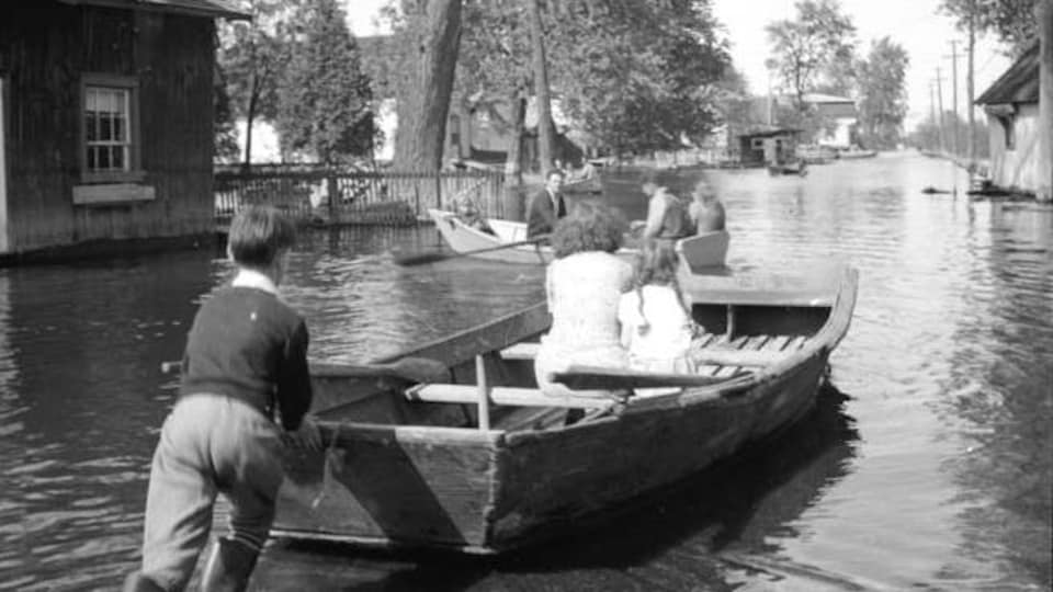 Une photo en noir et blanc illustre une barque qu'un garçon pousse dans une rue inondée.